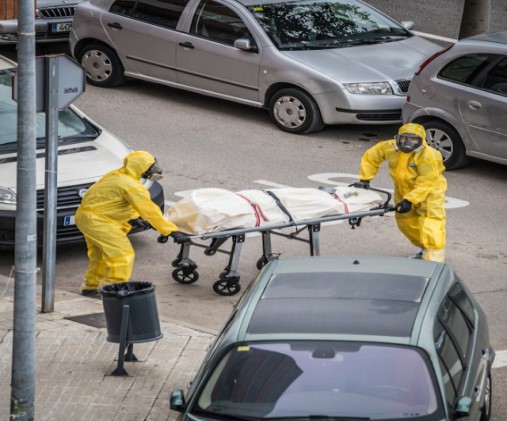 Dos hombres con mascarilla y vestidos de amarillo trasladan un cadáver en una camilla durante la pandemia de Covid.